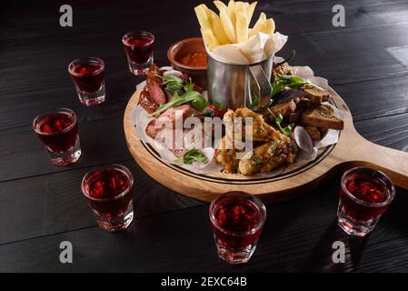 Assortiment d'en-cas : saucisses, ailes de poulet, frites, croûtons, sauce sur une assiette en bois sur une table en bois sombre. Alcool rouge en piles aroun Banque D'Images