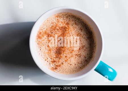 Couche plate dure et légère d'un latte de thé moussant recouvert de cannelle dans une tasse bleue sur une surface blanche. Banque D'Images