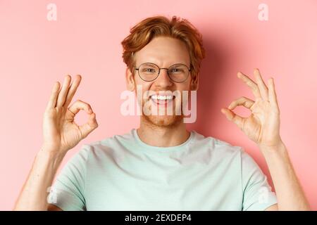 Portrait de tête d'un jeune homme attrayant avec des cheveux et une barbe rouges désordonnés, portant des lunettes, souriant avec des dents blanches et montrant des signes OK dans l'approbation Banque D'Images