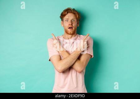 Homme nerveux avec des cheveux rouges épris indécise, bras croisés et pointant sur le côté, montrant deux variantes avec le visage perplexe, debout dans le t-shirt sur la menthe Banque D'Images