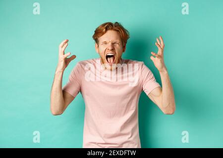 Un jeune redhead en colère criait affligé, se secouant les mains et se tenant au-dessus d'un fond turquoise Banque D'Images