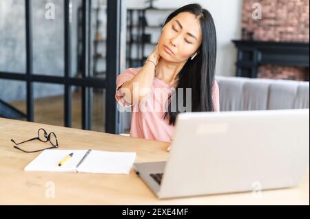 Femme asiatique fatiguée de travail en ligne sur un ordinateur portable massant son cou avec les yeux fermés, femme freelance sentir la fatigue assis au bureau à domicile Banque D'Images