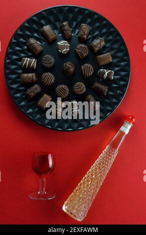 Diverses pralines au chocolat sur une assiette noire, un verre rouge et une bouteille de liqueur sur fond rouge Banque D'Images