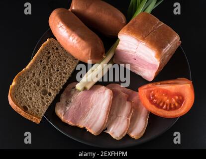 Aliments, oignons verts de saindoux, tomate, pain noir, saucisses, sur une plaque noire sur fond noir Banque D'Images