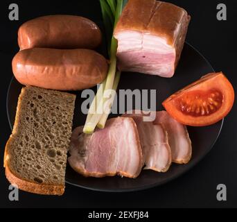 Aliments, oignons verts de saindoux, tomate, pain noir, saucisses, sur une plaque noire sur fond noir Banque D'Images