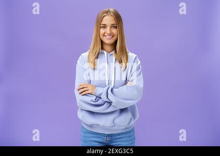 Portrait de charmante femme européenne charismatique avec la peau bronzée dedans un sweat à capuche tendance de grande taille se croisant les mains contre la poitrine en souriant largement appareil photo Banque D'Images