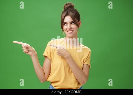 Coup de pouce de femme charmeur charismatique heureuse et insouciante dans t-shirt jaune pointant et regardant gauche enthousiaste et heureux souriant gaiement Banque D'Images