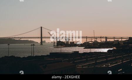 Coucher de soleil à Lisbonne, Portugal: Silhouettes du pont 25 de Abril et docks en arrière-plan, toits de maisons, une gare en premier plan, str Banque D'Images