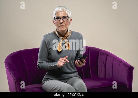 Une femme d'âge moyen de race blanche assise sur un canapé violet avec un téléphone et se confondre avec un fond blanc Banque D'Images