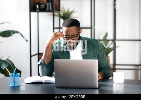 Homme d'affaires noir mécontent et multiracial ciblé, regard découragé sur l'ordinateur portable, message inattendu ou nouvelles, assis sur le lieu de travail Banque D'Images