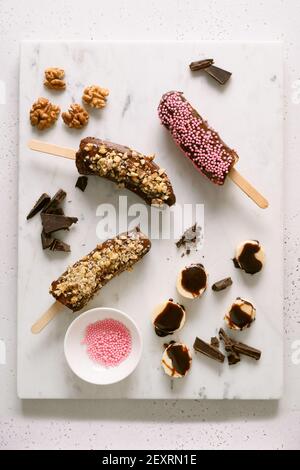 Sucettes glacées à la banane et au chocolat avec noix et saupoudrées roses. Des bonbons végétaliens faits maison faciles. Vue de dessus. Banque D'Images