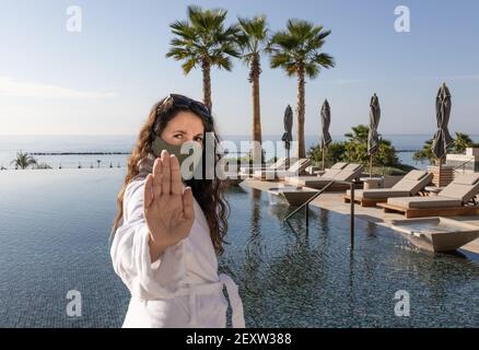 Jeune femme avec masque de visage montrant le signe d'arrêt à la caméra demandant de maintenir une distance de sécurité au luxe piscine du complexe Banque D'Images