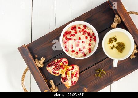 Petit déjeuner sain, flocons d'avoine avec grenade et tasse de thé vert, scène au-dessus sur une table en bois blanc. Banque D'Images