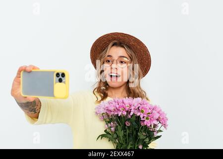 Belle jeune femme portant un chapeau de paille tenant un bouquet de fleurs roses fait selfie en utilisant son téléphone intelligent tout en se tenant sur fond blanc studio. Banque D'Images