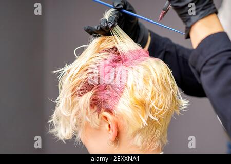 Gros plan sur les mains du coiffeur en appliquant du colorant rose sur celles de la femme cheveux blonds dans un salon de coiffure Banque D'Images