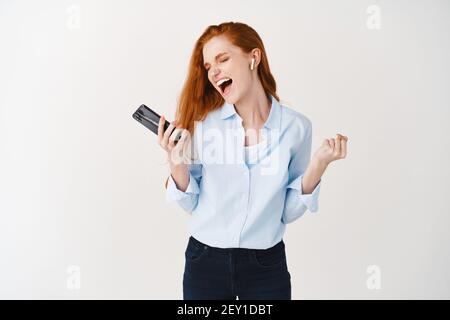 Jeune femme joyeuse avec de longs cheveux rouges chantant et écoutant de la musique dans un casque sans fil, s'amusant, tenant un smartphone, debout sur le blanc Banque D'Images