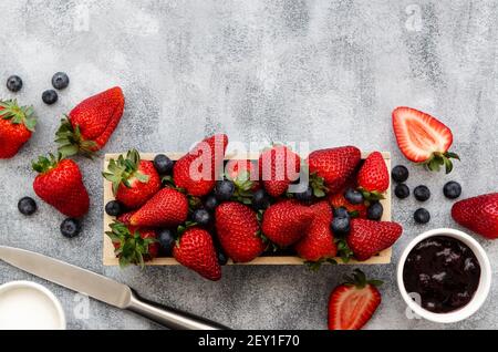 Fraises et bleuets dans une boîte rectangulaire en bois, confiture de fraises et de bleuets et sucre dans des bols blancs avec couteau, sur fond gris. Banque D'Images