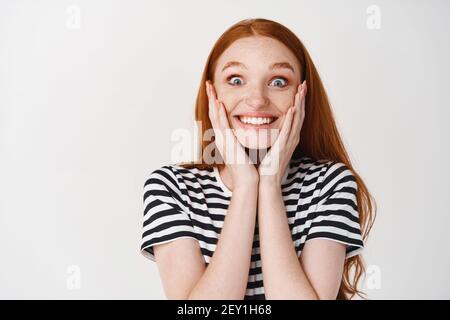 Image d'une jeune femme extrêmement heureuse et surprise avec des cheveux rouges, visage touchante et souriant émerveillé, recevoir un cadeau surprise, fond blanc Banque D'Images