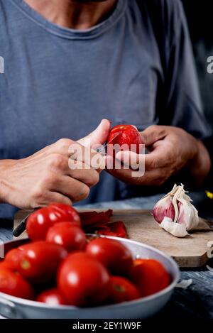 gros plan d'un jeune homme du caucase, portant un t-shirt gris, épluchant une tomate ébouillante avec un couteau sur une table de cuisine ou un comptoir à côté d'une tête de guar Banque D'Images