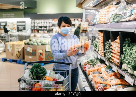 Épicerie. Femme d'âge moyen avec cheveux foncés courts en bleu masque de protection visage choisir l'achat de nourriture dans le supermarché et mettre dans le panier. Banque D'Images