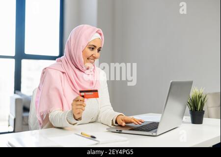 Une jeune femme musulmane heureuse portant le hijab et des vêtements décontractés intelligents tient une carte de débit et fait des commandes en ligne sur l'ordinateur portable assis dans un bureau contemporain, faire des achats, faire des transactions sur un compte bancaire Banque D'Images