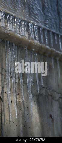 des glaçons pointus congelés suspendus à des tuyaux en métal industriel sur de l'eau grungie ont endommagé les ombres à glace du bâtiment en béton sur le mur extérieur en béton en arrière-plan Banque D'Images