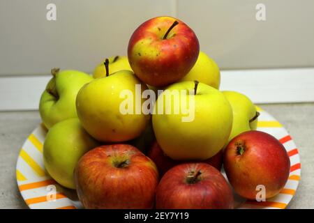 Tas de pommes dans une assiette sur la table. Les pommes mûres juteuses rouges et vertes se trouvent sur une lame sur une plaque blanche contre le fond d'un mur gris. Photo de stock avec vue sur le dessus des fruits Banque D'Images