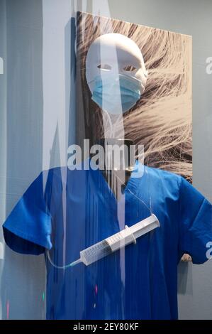 Sinister mannequin masqué vêtu de gommages chirurgicaux bleus, avec une seringue. Vitrine d'un salon de coiffure, pendant la pandémie du coronavirus. ROYAUME-UNI. Banque D'Images