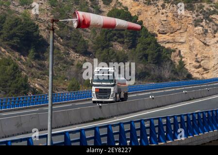 Camion-citerne avec des marchandises dangereuses circulant à travers un viaduc et une chaussette indique qu'il y a un vent fort. Banque D'Images