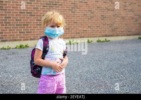 Petit enfant d'âge préscolaire dans le masque avec sac à dos pendant le virus corona près du bâtiment de l'école, aller à l'école ou à la maternelle. Premier jour à l'école concept. Banque D'Images