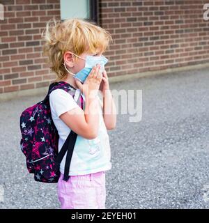 Petit enfant d'âge préscolaire dans le masque avec sac à dos pendant le virus corona près du bâtiment de l'école, aller à l'école ou à la maternelle. Premier jour à l'école concept. Banque D'Images