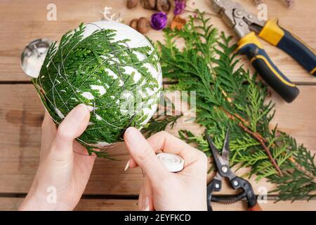 Fleuriste au travail: Femme montre comment faire boule de Noël avec sphère de styromousse et brindilles de thuja. Étape par étape, tutoriel. Banque D'Images