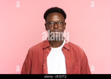 Homme afro-américain sérieux en lunettes porter une chemise rouge, regardant la caméra debout isolée sur rose Banque D'Images