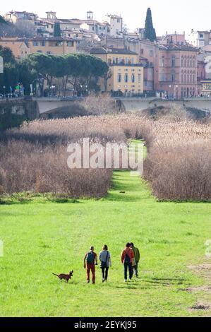 Florence, Italie - 2021, février 21 : un groupe d'amis avec un chien marchant sur un chemin herbeux le long de la rive de l'Arno. Paysage urbain inhabituel de Florence Banque D'Images
