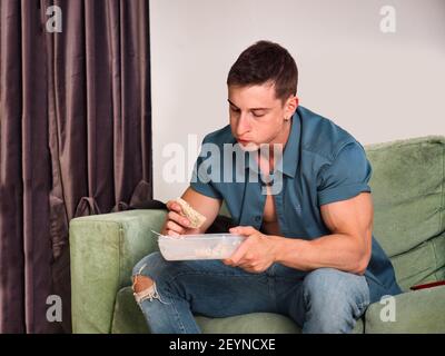 Un jeune homme musclé et affamé, qui ramasse la nourriture sur son canapé Banque D'Images