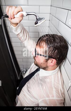 Homme en stress après le travail. Il se tient dans la salle de bains en lunettes et en vêtements, versant de l'eau courante sur lui-même à partir d'un arrosoir. Banque D'Images