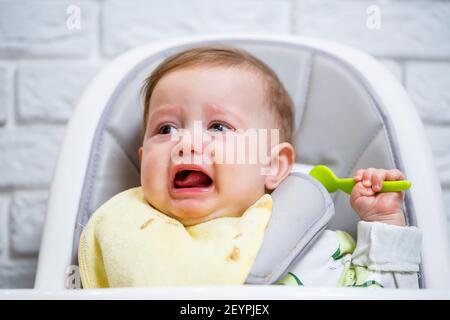 Un bébé souriant de neuf mois s'assoit à une table blanche dans une chaise haute et mange avec une cuillère dans un bol. Maman nourrit le bébé à partir d'une cuillère. Rétrogro flou Banque D'Images