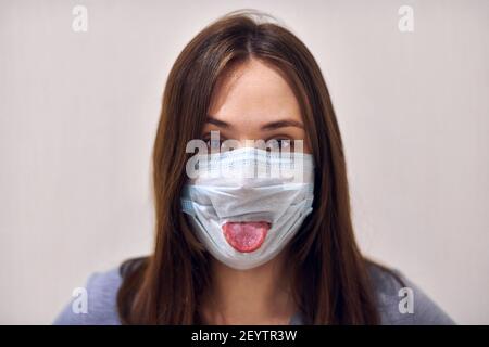 Portrait d'une femme drôle portant un masque médical et collant sa langue, gros plan. Concept de coronavirus fou. Banque D'Images