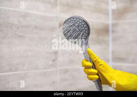 Main femelle dans gant de protection en caoutchouc jaune tenant la pomme de douche avec l'eau versante dans la salle de bains, espace de copie, mouvement flou. Banque D'Images
