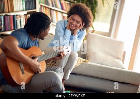 Une jeune femme aime le son de guitare joué par son mari dans une atmosphère détendue à la maison. Famille, ensemble, amour, musique Banque D'Images