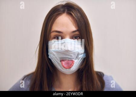 Portrait d'une femme drôle portant un masque médical et collant sa langue, gros plan. Concept de coronavirus fou. Banque D'Images