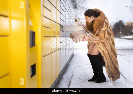 Une femme richement habillée ramasse un colis dans un lieu de cachette dans un casier à colis. Machine automatique pour l'envoi et la réception de lettres et de colis. Banque D'Images