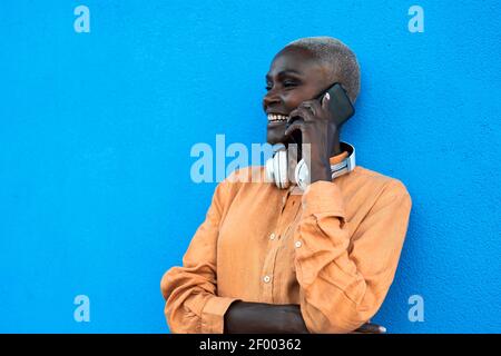 Une femme africaine qui fait un appel avec un smartphone mobile en étant debout sur fond bleu Banque D'Images