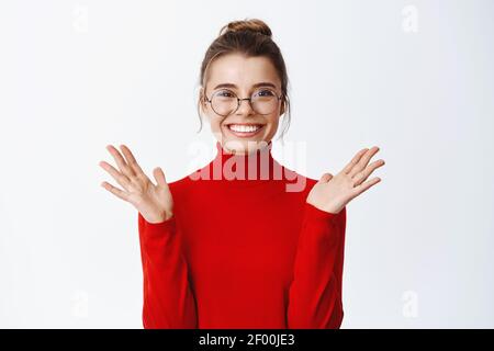 Portrait d'une jeune femme PDG en lunettes et chandail rouge, levant les mains de bonnes nouvelles joyeuses, souriante excitée, debout contre le blanc Banque D'Images