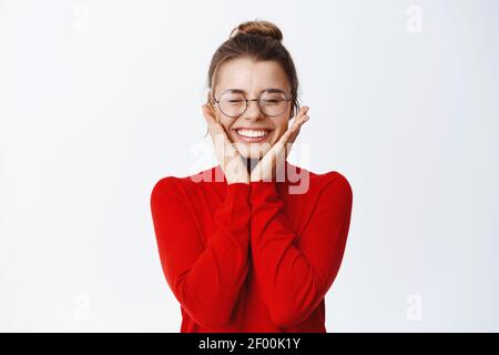 Portrait de blonde gaie femme d'affaires en lunettes se réjouissant, les yeux proches et touchant les joues, atteindre le but et sauter de joie excessive, se tenant sur blanc Banque D'Images