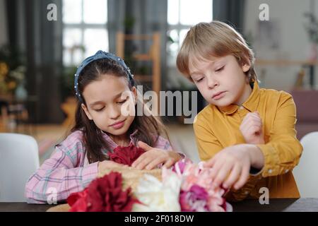 Mignon petit garçon et fille en tenue décontractée assis près d'une table devant l'appareil photo et mettant des fleurs faites à la main de couleurs blanc, rose et rouge sur le chapeau Banque D'Images