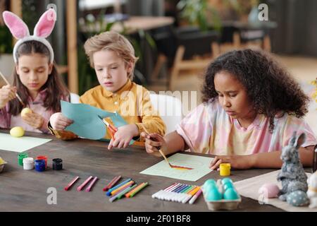 Groupe de trois petits enfants adorables et amicaux de diverses origines ethniques assis près d'une table dans un environnement familial et en préparant des cadeaux Pâques Banque D'Images