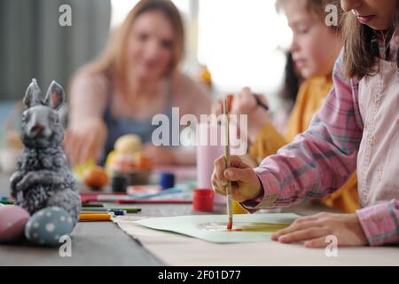 Petite fille sérieuse en vêtements décontractés peinture gros oeuf de Pâques papier assis sur une table en bois contre ses amis se préparant pour les vacances Banque D'Images