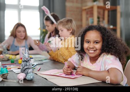 Joyeux mélange-race petite fille peinture grand oeuf de Pâques sur papier tout en étant assis sur une table en bois contre ses amis se préparant pour vacances Banque D'Images