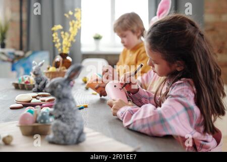 Petite fille sérieuse mignon avec highliget dessin visage de Pâques lapin sur papier roulé en étant assis près de la table garçon d'âge élémentaire Banque D'Images